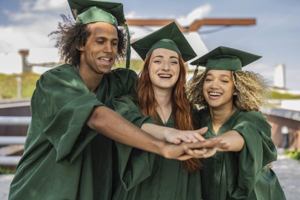three friends in graduation gowns posing to camera on college campus, joining hands, laughing, happy graduation day, close-up portrait